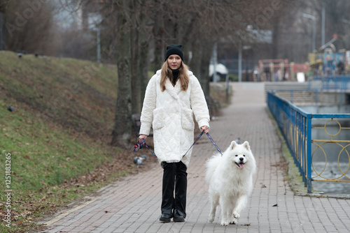 A woman in a cozy white coat walks her fluffy white samoyed dog along a serene pathway. The riverbank in the background adds to the tranquil atmosphere of this cool day