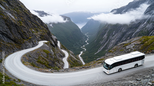 A white bus travels along a narrow winding road carved into steep mountainsides, overlooking a lush green valley and river below in a dramatic fjord landscape under overcast skies