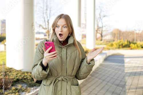 Smiling woman using mobile phone app, playing game, shopping online outdoors. Happy woman celebrating victory in lottery. Amazed hipster wondered with win in online auction betting on website photo