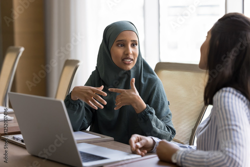 Arabian woman dressed in hijab discuss project updates with Latina colleague, delegating tasks and responsibilities, gather for budget allocation and financial planning, provide consultation to client