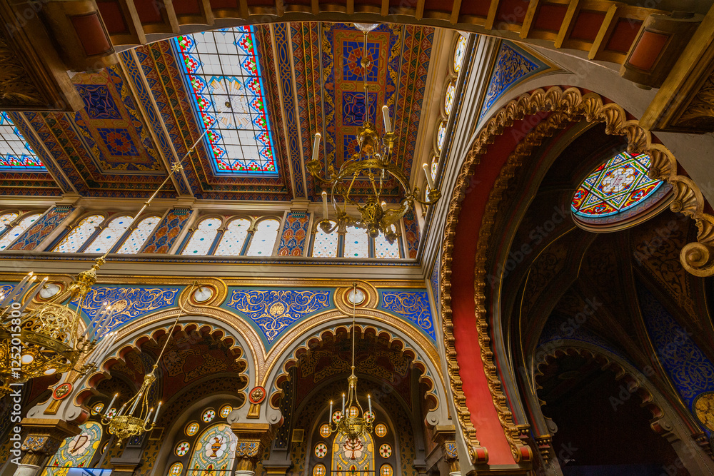 Fototapeta premium A captivating interior view of a synagogue showcasing its ornate ceiling with geometric patterns, arched windows, and a portion of an elaborate chandelier, all bathed in soft, natural light.
