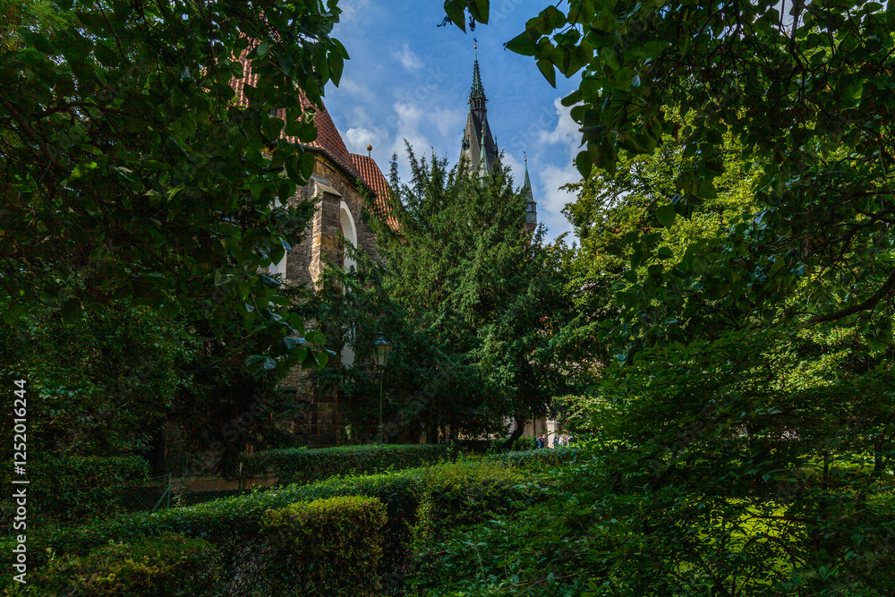 A picturesque scene of a historic building with a tall spire and red roof, partially obscured by lush green trees and foliage, seen on a bright day with blue sky and white clouds