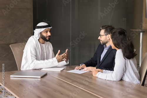 Latin couple listening Arabian male real estate agent, property seller dressed in dishdasha makes speech, explaining contract details, selling company services to European spouses at meeting in office photo