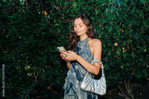 A fashionable woman checks her smartphone while smiling, standing against lush green bushes, dressed in a trendy summer outfit