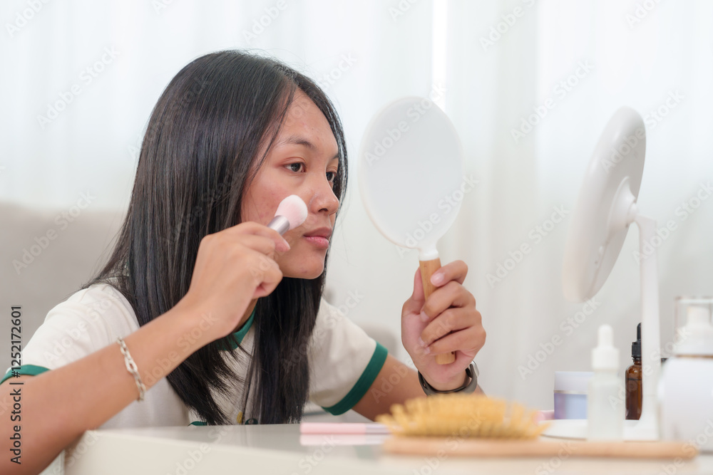 Asian young woman with long black hair applying makeup with soft brush while looking into handheld mirror. Natural skin texture visible. Beauty products, brushes, and skincare items neatly arranged Asian young woman with long black hair applying makeup with soft brush while looking into handheld mirror. Natural skin texture visible. Beauty products, brushes, and skincare items neatly arranged