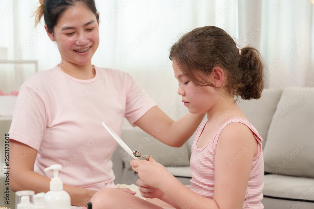 Caucasian girl learning makeup with Asian mom. Little girl carefully using eyelash curler while mother watches with smile. Beauty products and brushes arranged on table in cozy home environment. Caucasian girl learning makeup with Asian mom. Little girl carefully using eyelash curler while mother watches with smile. Beauty products and brushes arranged on table in cozy home environment.
