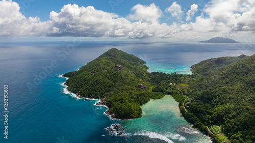Expansive azure ocean with a green island featuring rocky shores and dense forest vegetation. Seychelles, Mahe.
