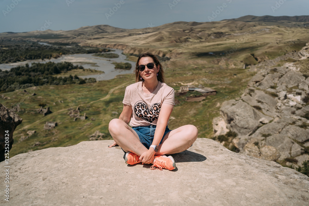 Young woman sitting cross-legged on a cliff with a view of the valley on a sunny day. Woman Sitting in Lotus Pose on a Rock with Scenic Valley View Young woman sitting cross-legged on a cliff with a view of the valley on a sunny day. Woman Sitting in Lotus Pose on a Rock with Scenic Valley View