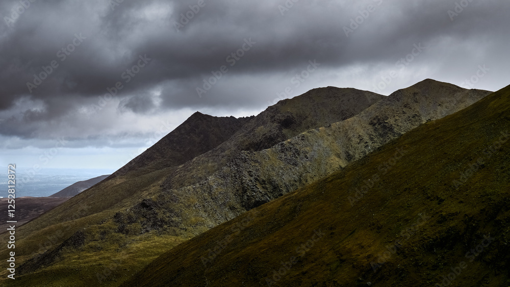 Jagged mountain ridges under a dramatic, cloudy sky, visible lower terrain with some greenery. Jagged mountain ridges under a dramatic, cloudy sky, visible lower terrain with some greenery.