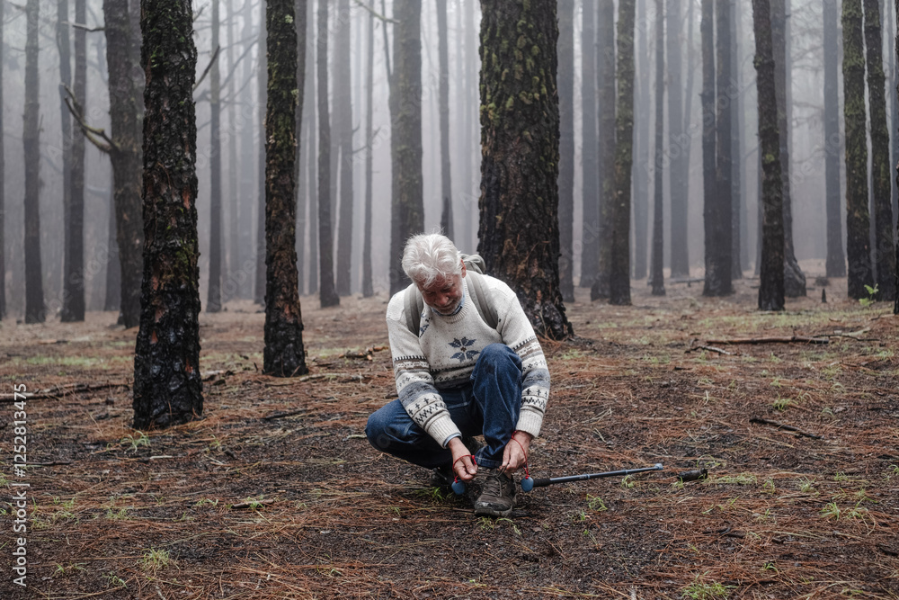 Fototapeta premium Active happy senior retired man with backpack walking in mountain forest on a foggy day stops to tie his shoes. Bare trees on backgrounds