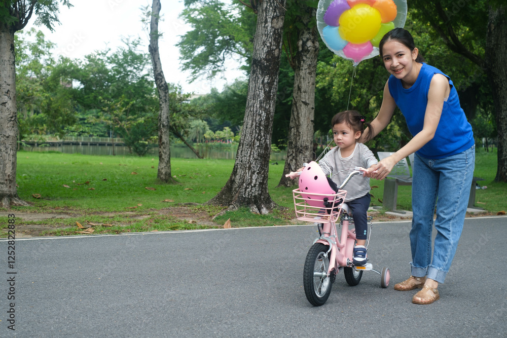 A joyful scene of a woman helping a young child ride a pink bicycle, surrounded by colorful balloons in a sunny park. A joyful scene of a woman helping a young child ride a pink bicycle, surrounded by colorful balloons in a sunny park.
