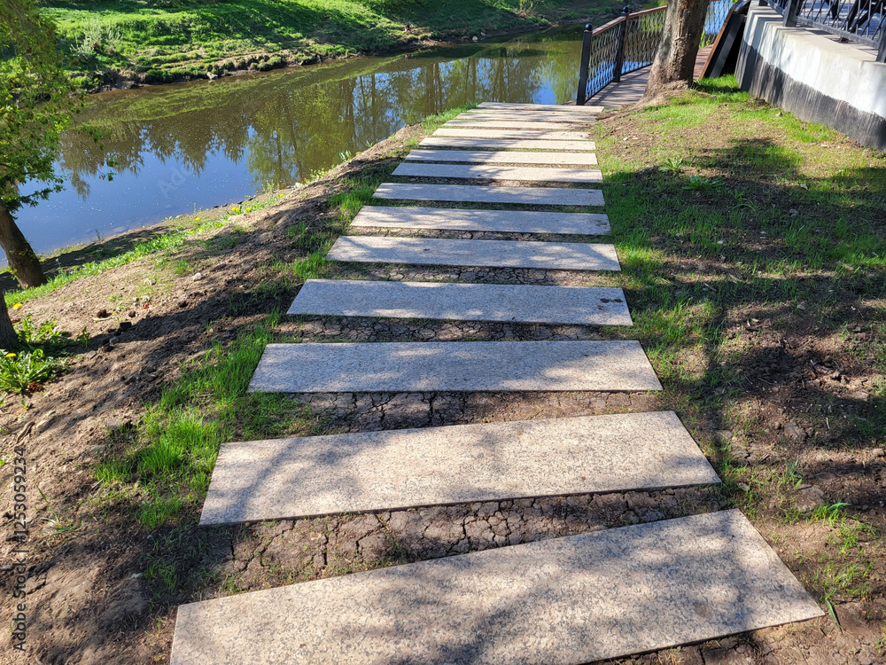 Obraz premium A path of concrete rectangles is laid along a shady spring lawn along the river. The concept of landscaping and landscape design