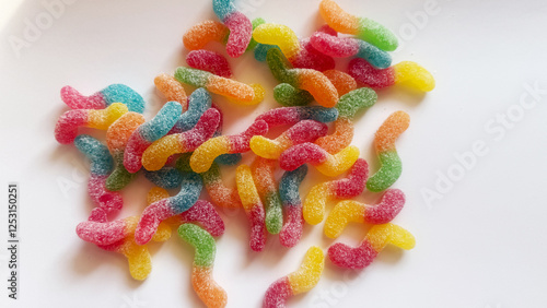 A vibrant assortment of sugar-coated gummy worms in various bright colors, including red, green, yellow, blue, and orange, placed in a transparent glass bowl. The candies have a soft, chewy texture