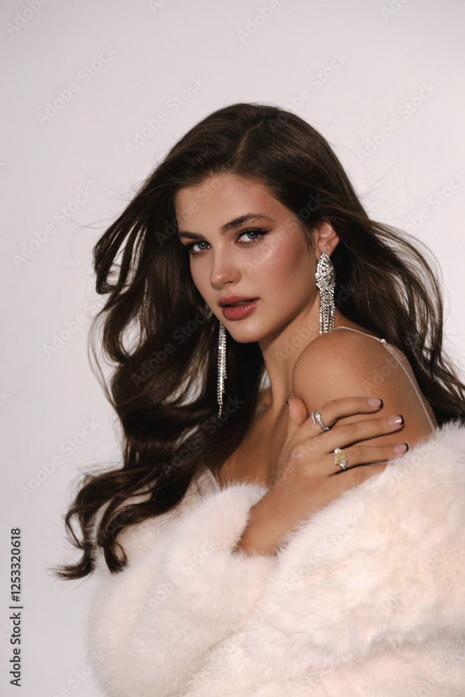 Naklejka premium beautiful woman with dark hair in luxurious white fur coat and jewelry posing in studio