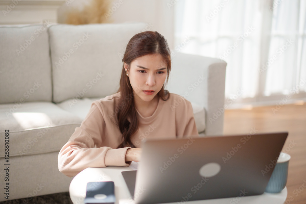 Asian woman working from home with a serious and focused expression, seated at a coffee table in a living room while using her laptop. Asian woman working from home with a serious and focused expression, seated at a coffee table in a living room while using her laptop.