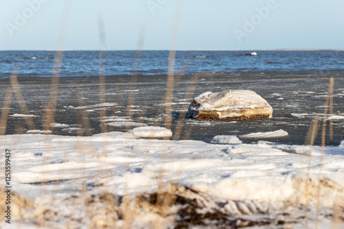 Icy seashore with scattered snow and a frosty boulder. A serene winter scene featuring a frozen seashore, icy surface, scattered snow patches, and a frost-covered boulder under a clear blue sky.