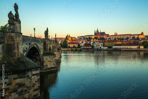 A stone bridge with statues lining its edge spans a wide river, offering a distant view of a cityscape with red-roofed buildings and a large complex under a clear sky at twilight.