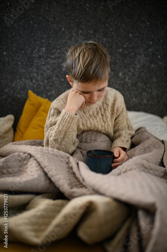 A boy with a cold, fever, and cough drinks ginger tea with lemon in bed. The soothing tea helps him feel better, offering comfort and warmth during his recovery.