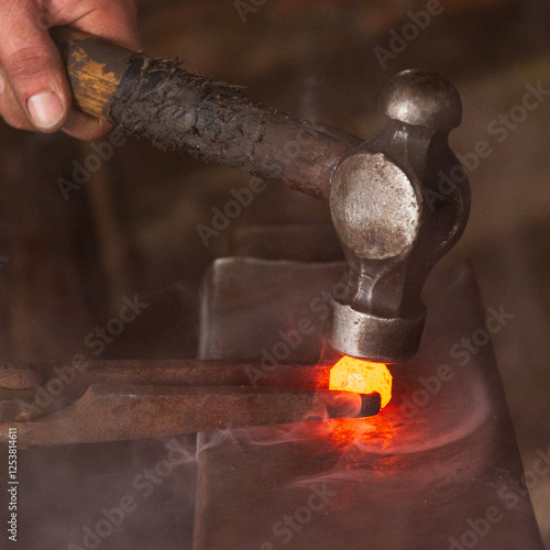 A hammer being used to shape a sphere of red hot steel held by tongs on an anvil in a forge