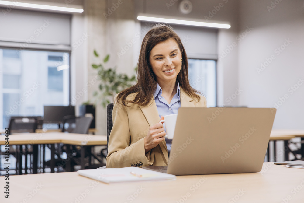 Smiling businesswoman enjoying a cup of coffee while working on a laptop in a bright, modern office space filled with natural light Smiling businesswoman enjoying a cup of coffee while working on a laptop in a bright, modern office space filled with natural light