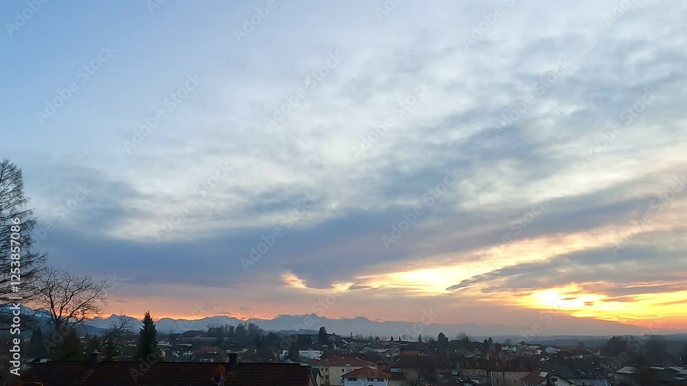 breathtaking sunset over a picturesque European town with the silhouette of distant mountains on the horizon. The sky is painted in shades of orange, yellow, and blue, with soft clouds stretching acro breathtaking sunset over a picturesque European town with the silhouette of distant mountains on the horizon. The sky is painted in shades of orange, yellow, and blue, with soft clouds stretching acro