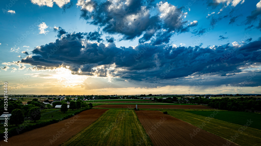 Obraz premium A vibrant sunset casts warm colors over expansive farmland, with lush green fields and patches of clouds creating a picturesque sky. The serene landscape reflects the tranquility of rural life.