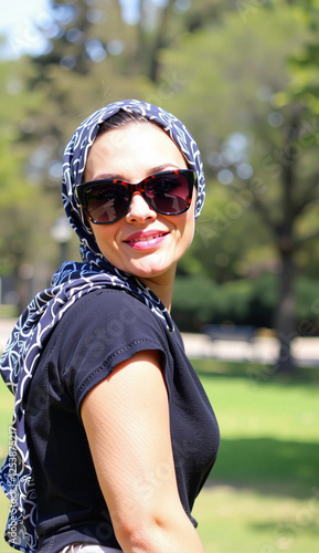 Stylish woman with sunglasses and scarf smiling in park during sunny day
