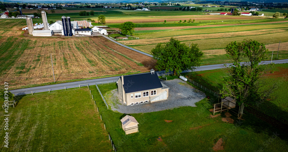 Fototapeta premium An Amish one room school house sits on a gravel area, surrounded by expansive green fields, barns, and distant structures under a bright blue sky.
