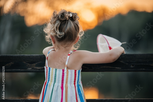 Obraz na plátně A small girl about 5 years old standing with her back to the camera, looking at a warm sunset horizon
