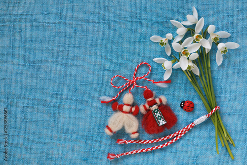 Bouquet of snowdrops and a red and white martenitsa bracelet, figures of a boy and a girl on a blue fabric background. Postcard for the holiday of March 1, Martisor, Baba Marta