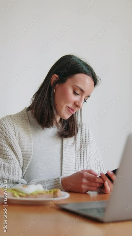 Vertical video, Smiling woman working remotely. She is relaxing on her couch using her laptop and phone. Vertical video, Smiling woman working remotely. She is relaxing on her couch using her laptop and phone.