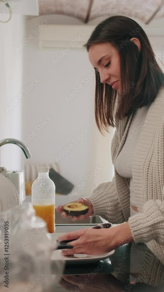 Vertical video, A young woman is happily preparing an avocado for breakfast in her modern kitchen. Vertical video, A young woman is happily preparing an avocado for breakfast in her modern kitchen.