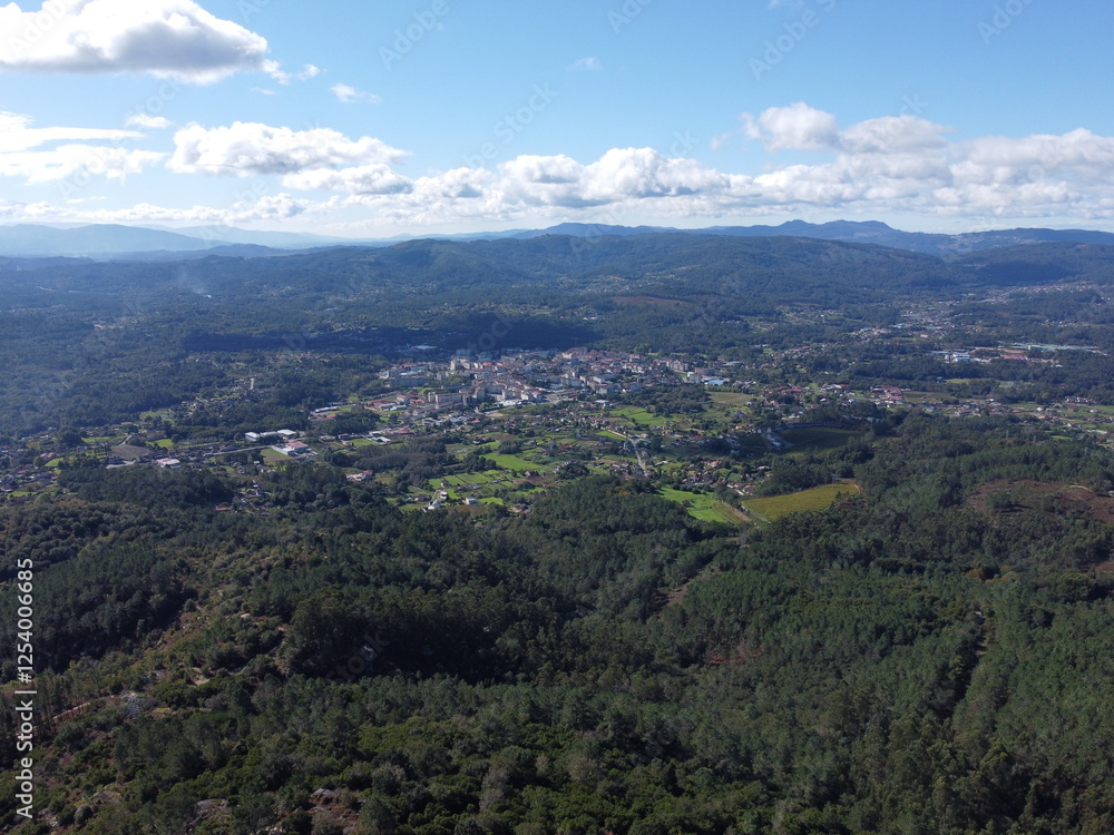 Aerial view of Ponteareas, with lush green forests, agricultural fields, and the town spread out across the valley, surrounded by rolling hills under a clear blue sky with fluffy clouds, showcasing th Aerial view of Ponteareas, with lush green forests, agricultural fields, and the town spread out across the valley, surrounded by rolling hills under a clear blue sky with fluffy clouds, showcasing th