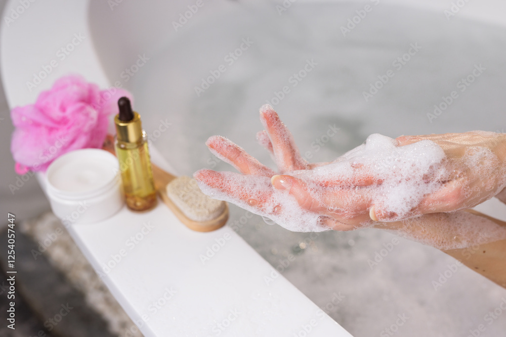 Fototapeta premium Relaxing spa moment. Woman washing hands with foamy soap near a bathtub. Skincare essentials like cream, oil, brush, and loofah on the side. Self-care, hygiene, and wellness concept