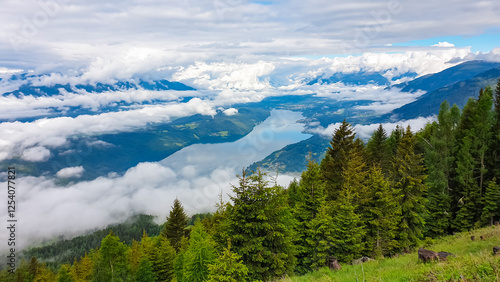 From summit of Mirnock, breathtaking panorama unfolds, revealing Lake Millstatt in Carinthia, Austria. Nestled amidst rolling hills and valleys cloaked in sea of clouds. Evergreen forests blanket photo