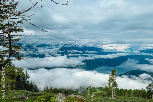 Majestic Lake Millstatt rests serenely below, viewed from Mirnock, Austria. Clouds embrace mountains, creating dreamy, elevated landscape framed by lush trees and vegetation. Alpine lush green pasture photo