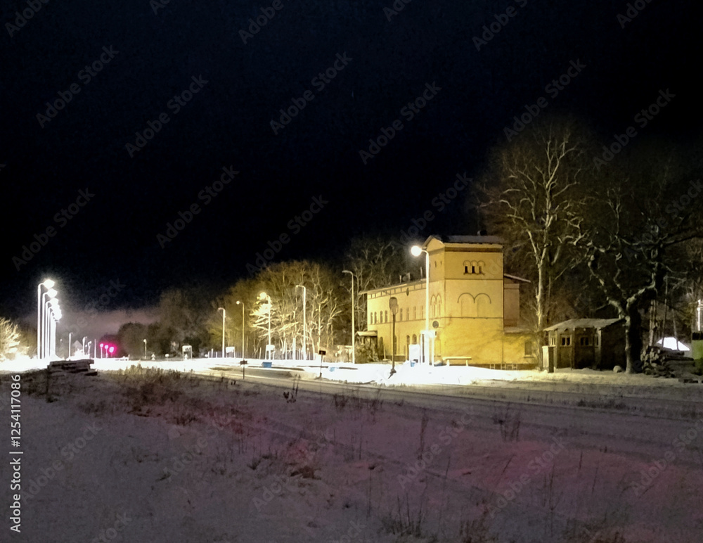 Mała prowincjonalna stacja kolejowa w zimową noc. Perony oświetlają lampy elektryczne. Dachy i ziemię pokrywa śnieg.