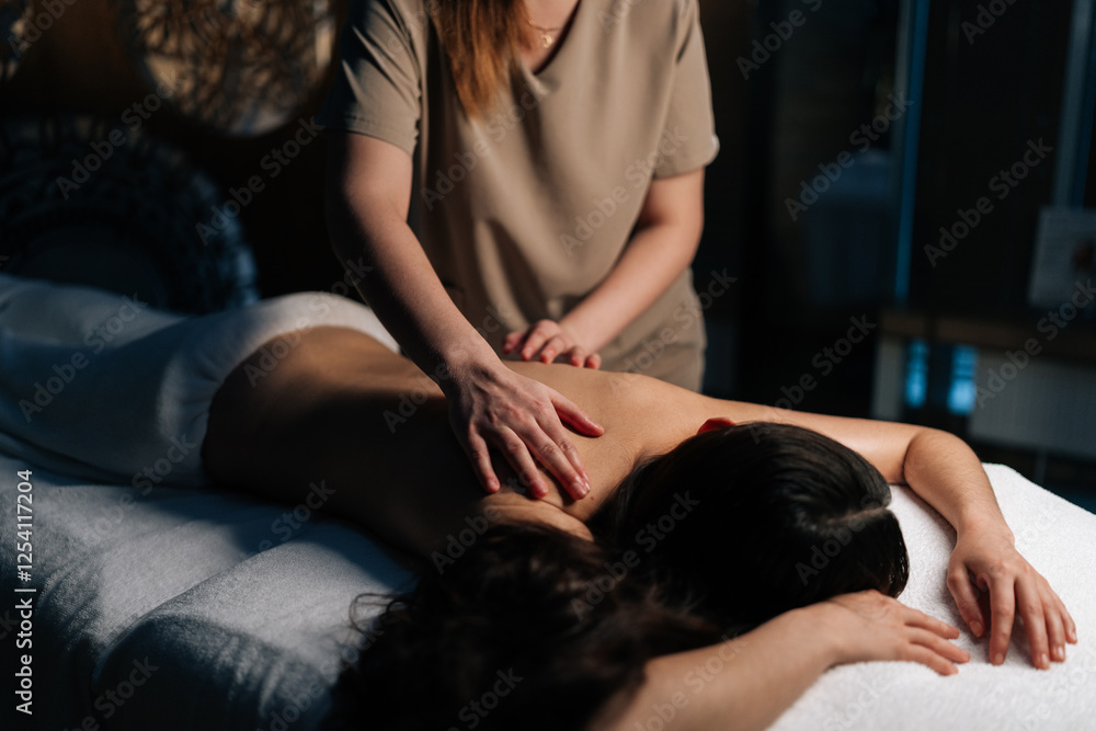 Obraz premium Closeup of unrecognizable masseuse applying massage oil back of young woman lying on massage table, providing relaxing and therapeutic body treatment in spa salon with dark interior.