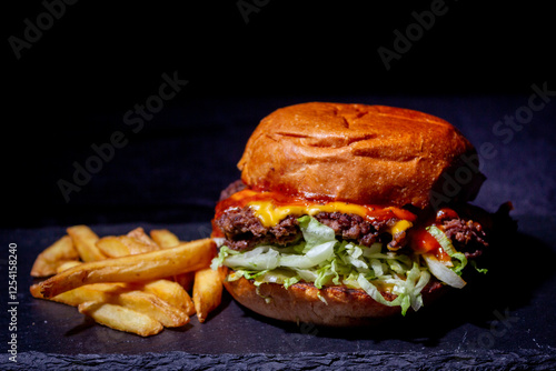Very delicious burger , with juicy meat, salad, a slice of  cheese and ketchup, with a golden crust and golden fries on the side on the black stone table