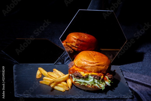 Delicious burger with golden crust in mirror reflection with juicy meat, salad, melted cheese slice and ketchup, next to golden fries on black stone plate and black velvet
