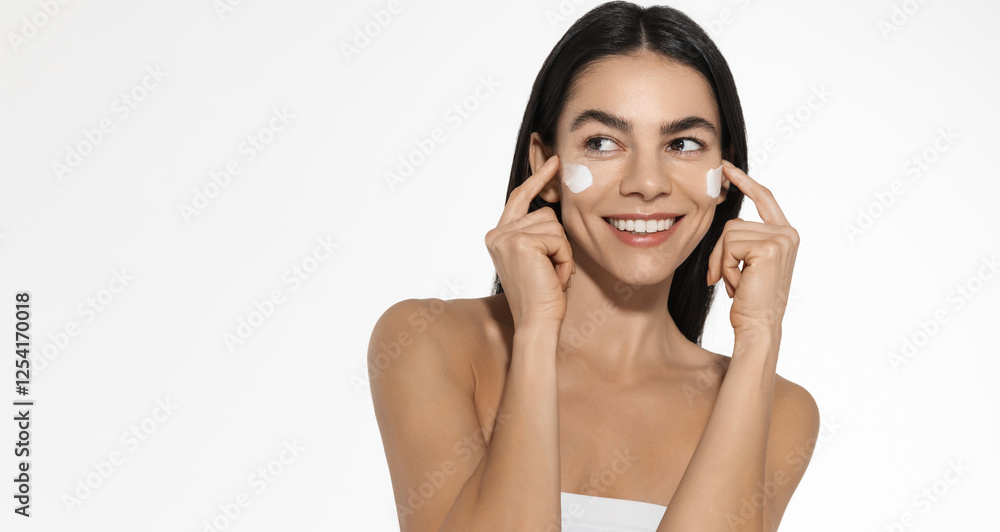 Female model with perfect skin and white smile, applies moisturising cream on her cheeks, using treatment from blemishes, anti-aging product, looking away while standing over white background Female model with perfect skin and white smile, applies moisturising cream on her cheeks, using treatment from blemishes, anti-aging product, looking away while standing over white background