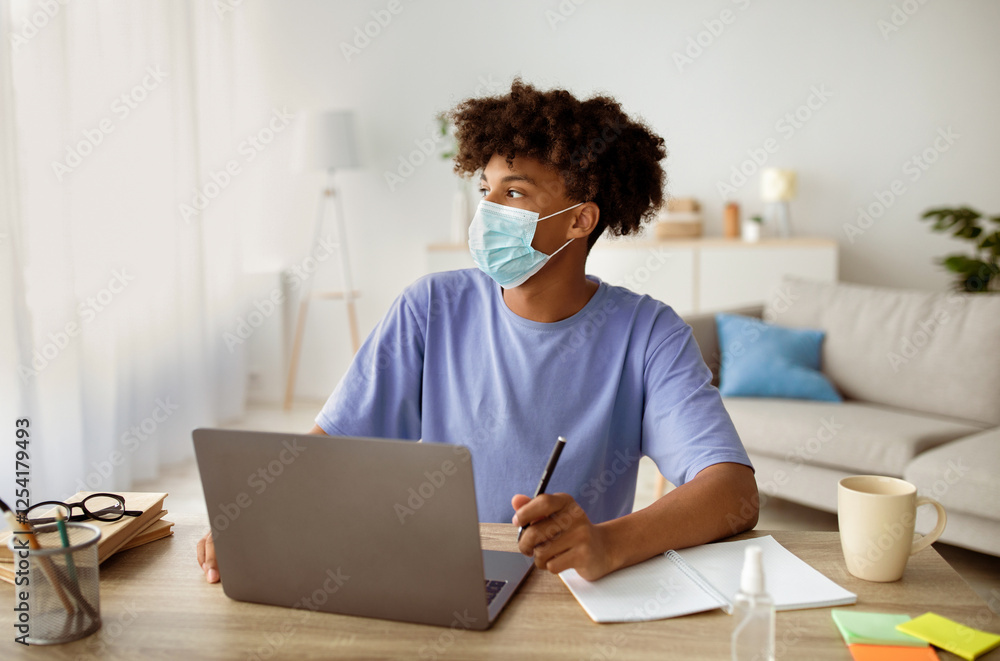 Black teenage student in face mask sitting at desk with laptop, taking notes in copybook, studying online from home during covid isolation. African American adolescent on homeschooling Black teenage student in face mask sitting at desk with laptop, taking notes in copybook, studying online from home during covid isolation. African American adolescent on homeschooling