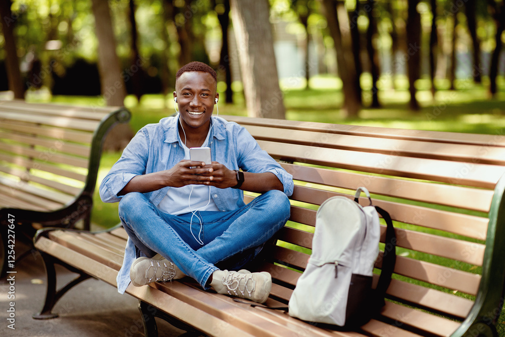 Obraz premium Cool black guy in earphones listening to music online from his smartphone on bench at summer park, empty space