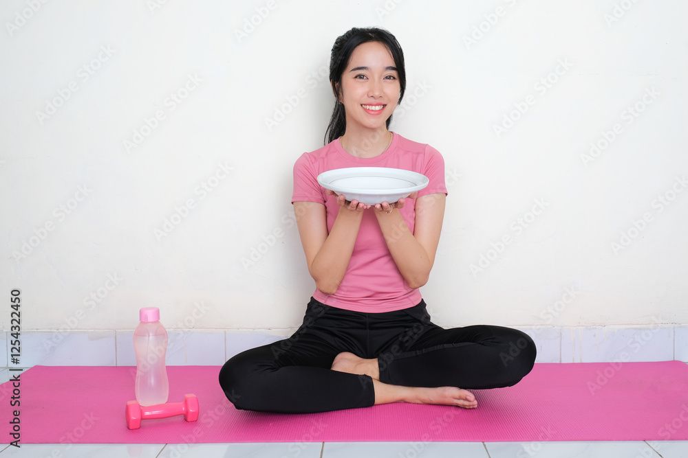 A woman in sportswear sitting on yoga mat holding empty dining plate