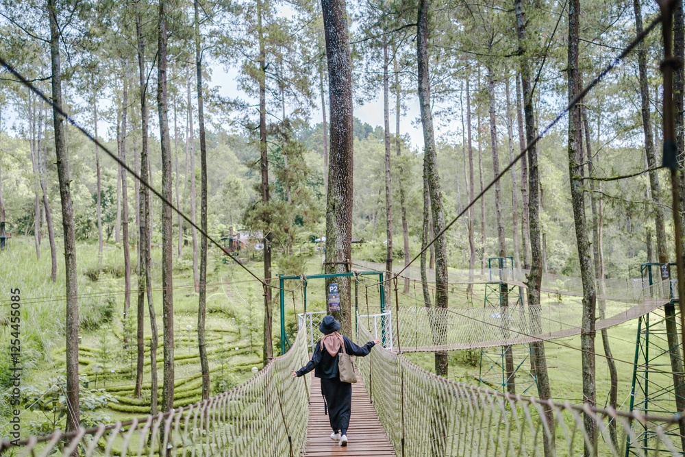A traveler wearing a hijab walks across a wooden suspension bridge in the middle of a lush pine forest. The scene exudes adventure, nature exploration, and eco-tourism vibes. A traveler wearing a hijab walks across a wooden suspension bridge in the middle of a lush pine forest. The scene exudes adventure, nature exploration, and eco-tourism vibes.