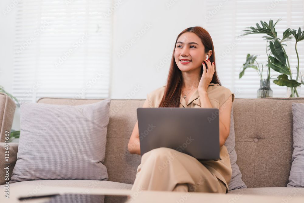 Woman sitting on sofa with laptop, smiling while using wireless earbuds in bright living room. Woman sitting on sofa with laptop, smiling while using wireless earbuds in bright living room.