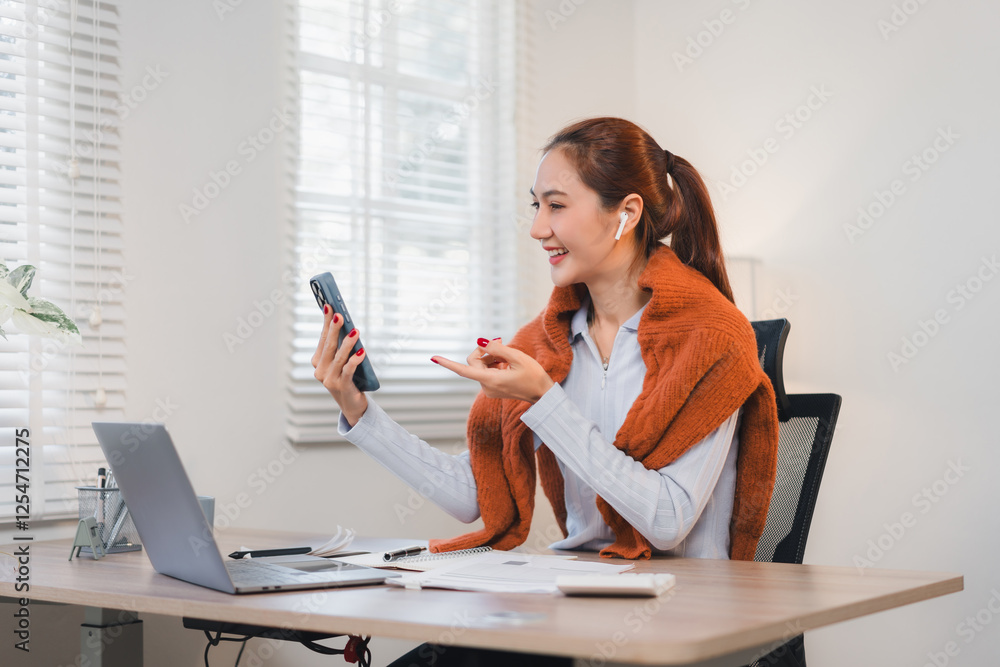 Obraz premium Woman in home office smiling while using smartphone and wearing wireless earbuds.