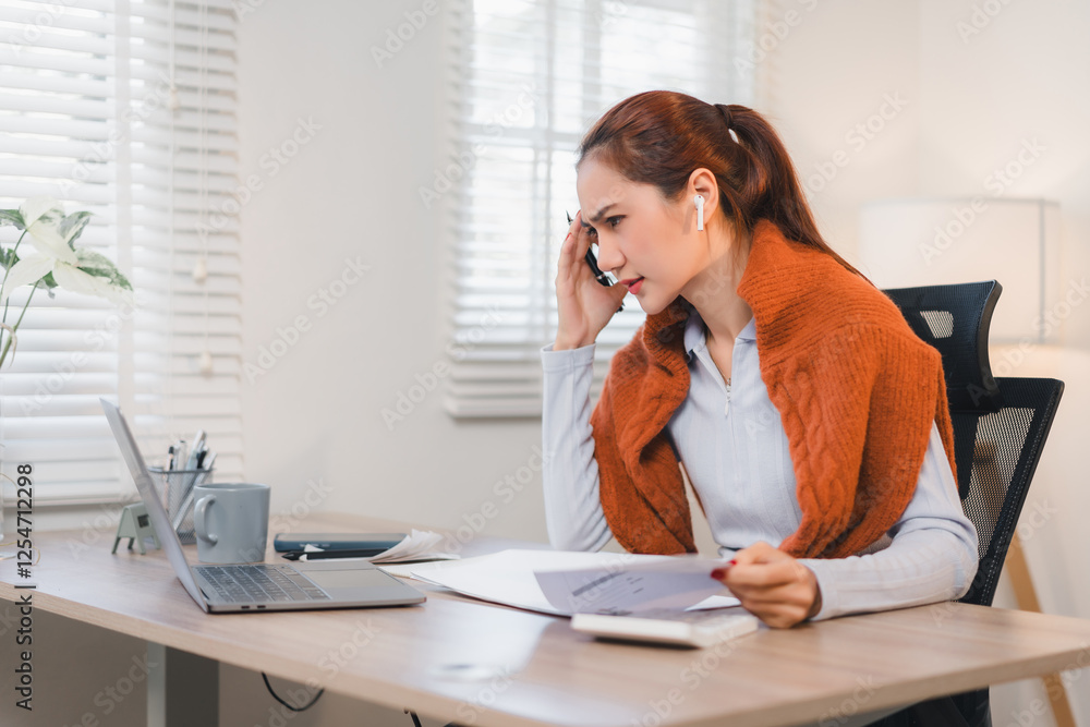 Obraz premium Woman working from home, wearing earphones, focused on laptop while talking on phone.
