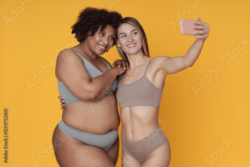 Two women are smiling while taking a selfie with a smartphone against a vibrant yellow background. They are embracing each other creating a joyful and friendly atmosphere photo