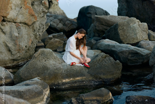 Woman in white dress sitting on ocean rocks with red shoes, gazing pensively at the horizon photo